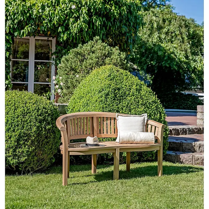 Teak Gartenbank Varadero Bananenbank 160 cm Massivholz wetterfest edel -  - Gebogene Gartenbank aus Naturholz mit Kissen und Hut, in einem sonnigen grünen Garten.