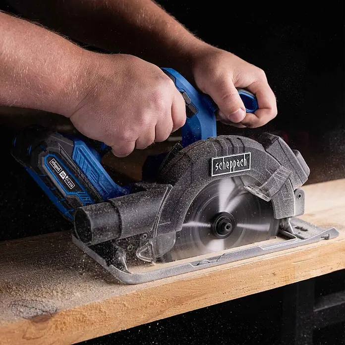Scheppach IXES 20V Akku-Handkreissäge C-HCS165-X - 20 V, Durchmesser Sägeblatt: 165 mm - Blaue und schwarze Scheppach 20V IXES Akku-Kreissäge schneidet ein Holzbrett, von Händen gehalten, mit Sägespänen.