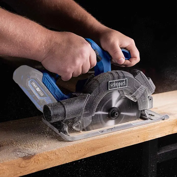 Scheppach IXES 20V Akku-Handkreissäge C-HCS165-X - 20 V, Durchmesser Sägeblatt: 165 mm - Akkubetriebene Scheppach Handkreissäge (blau/schwarz) schneidet Holz, Hände bedienen sie, Späne fallen.