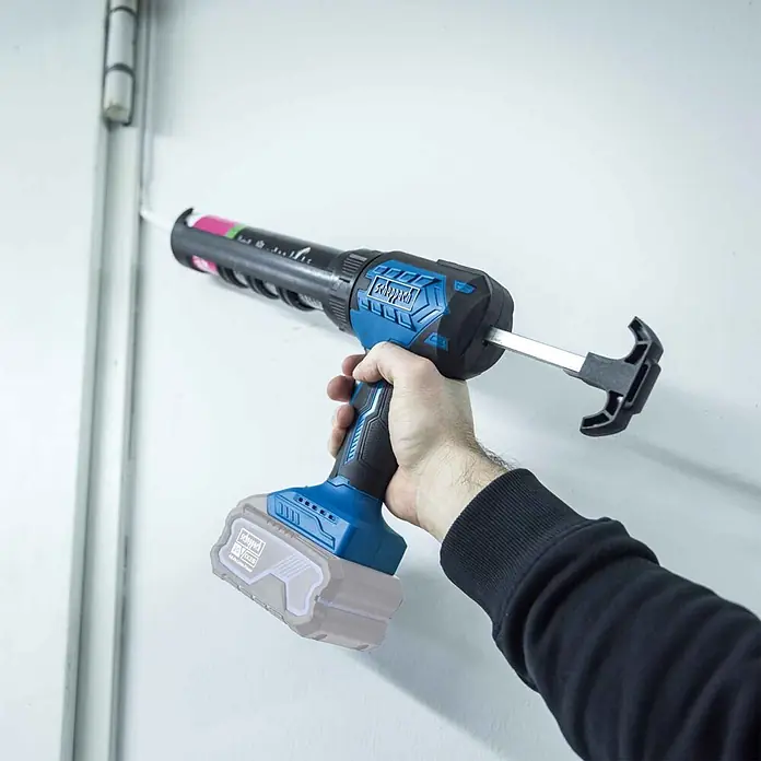 Scheppach IXES 20V Akku-Kartuschenpresse C-SEG310-X - 20 V, Ohne Akku - Eine Hand hält eine blau-schwarze Akku-Kartuschenpresse und trägt Dichtmittel auf eine weiße Oberfläche neben einem Metallrahmen auf.