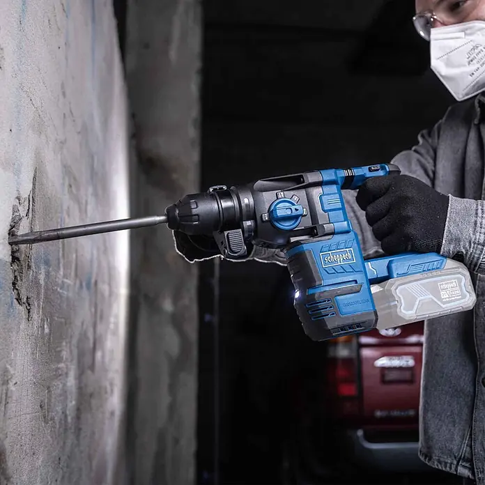 Scheppach IXES 20V Akku-Kombihammer - 20 V, Ohne Akku, 2,8 J - Person mit Maske und Handschuhen nutzt einen blau-schwarzen Scheppach Akku-Bohrhammer auf einer Betonwand.