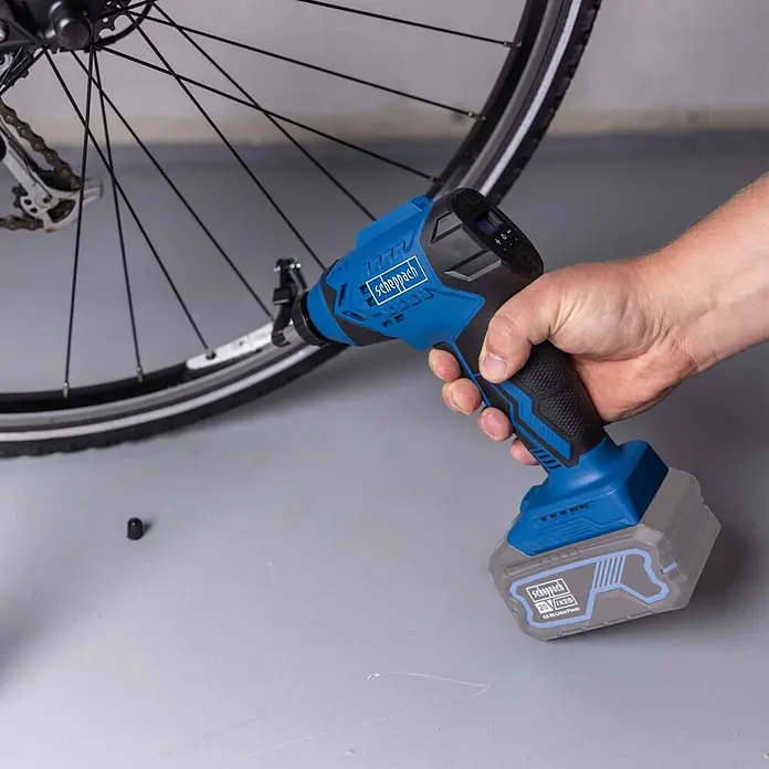 Scheppach IXES 20V Akku-Luftpumpe C-AC20-X - Max. Druck: 7 bar - Ein blauer und schwarzer scheppach Akku-Luftkompressor, von einer Hand gehalten, zum Aufpumpen an ein Fahrradreifenventil angeschlossen.