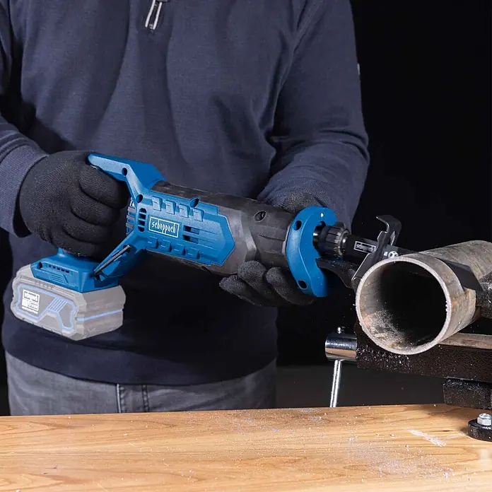 Scheppach IXES 20V Akku-Säbelsäge C-RS100-X - 20 V, Hublänge: 20 mm - Person in Handschuhen schneidet mit blauer und schwarzer Akku-Säbelsäge ein Metallrohr, das in einem Schraubstock auf einem Holztisch befestigt ist.