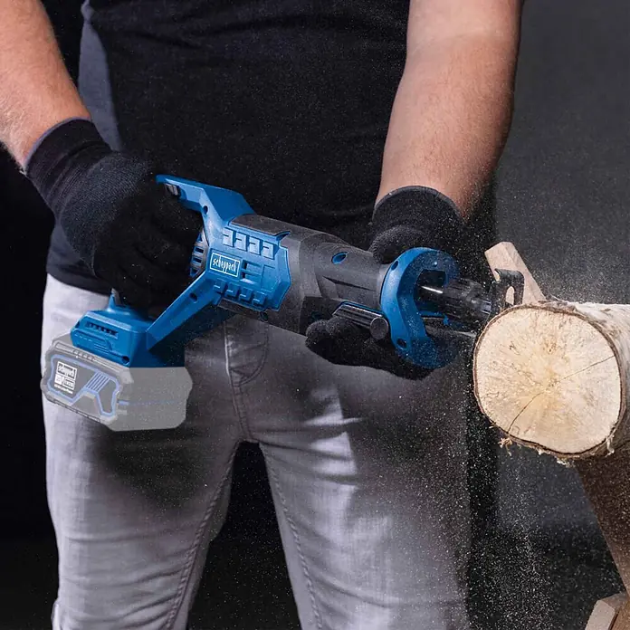 Scheppach IXES 20V Akku-Säbelsäge BC-RS250-X - 20 V, Ohne Akku, Hublänge: 28 mm - Person schneidet mit einer blauen und schwarzen Akku-Säbelsäge ein Holzstück, Sägespäne fliegen.