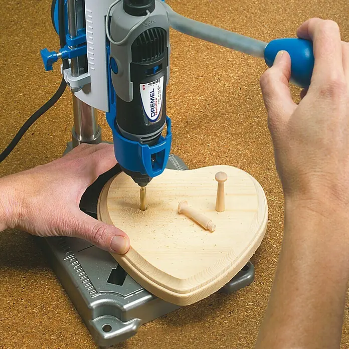 Dremel Workstation 220 - Passend für: Dremel Multifunktionsgeräte, Hub: 50 mm - Nahaufnahme einer Bohrmaschine, die ein Loch in ein hölzernes Herz bohrt, wobei der Fokus auf dem Dremel-Werkzeug und dem Werkstück liegt.
