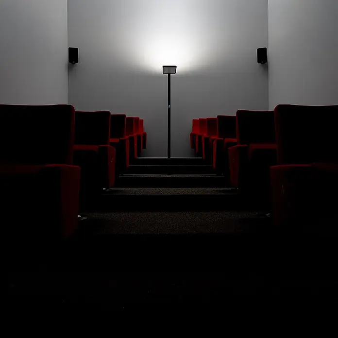 Luxula LED-Stehleuchte Stehlampe Up/Down Bürolampe Weiß 100W 7000LM 4000K -  - Eine schlanke schwarze Stehleuchte mit leuchtendem Kopf, die in einem dunklen Kinosaal mit roten Sitzen Umgebungslicht spendet.