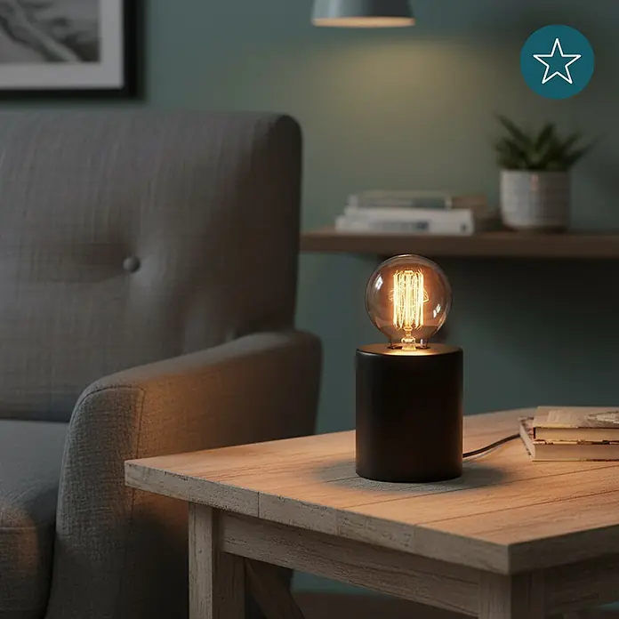Luxula Deko-Tischleuchte E27 Fassung Modern Design Metall Nachttischleuchte -  - Eine schwarze zylindrische Tischleuchte mit glühender Edison-Glühbirne steht auf einem hellen Holztisch neben einem grauen Sessel.
