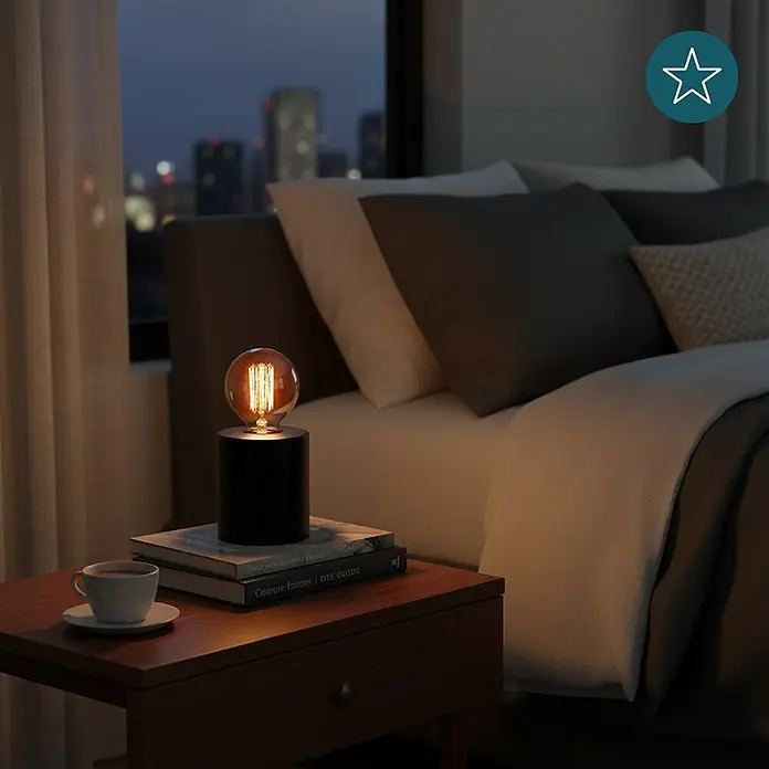 Luxula Deko-Tischleuchte E27 Fassung Modern Design Metall Nachttischleuchte -  - Eine schwarze zylindrische Nachttischlampe mit leuchtender Edison-Glühbirne steht auf Büchern auf einem Nachttisch neben einem Bett und beleuchtet das warme Schlafzimmer.