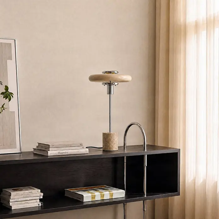 Lindby Tischlampe in beige, Nachttischlampe -  - Eine moderne Tischlampe mit hellbeigem Schirm, Chromschaft und Travertin-ähnlichem Sockel steht auf einem dunklen Regal.