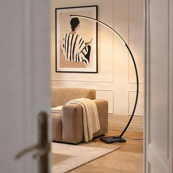 Lindby LED Stehlampe in schwarz, Bogenlampe Wohnzimmer -  - Eine schwarze, gebogene Stehlampe mit LED-Streifen und Marmorsockel, die Sofa und Kunstwerk in einem gemütlichen Raum beleuchtet.