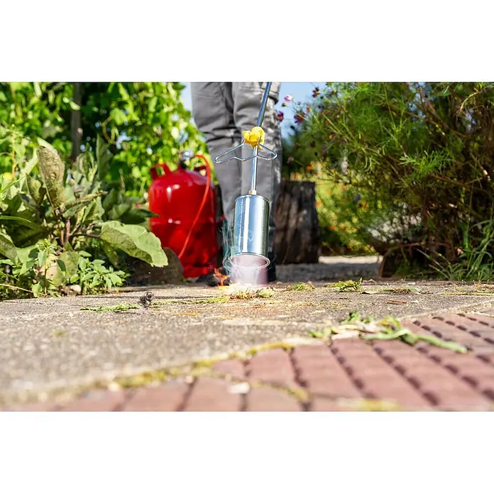 Gloria Unkrautvernichter Thermoflamm Professional - Flammtemperatur: 1.000 °C - Silberner Unkrautbrenner mit gelbem Griff, an rotem Gastank, brennt Unkraut auf einem gepflasterten Gartenweg ab.