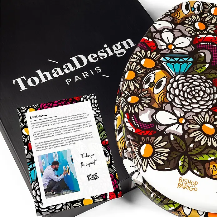 Tohaa Design WC-Sitz BirdyFlowers - Bishop Parigo -  - Schwarze TohaaDesign-Box mit einem runden, farbenfrohen Produkt, verziert mit einem einzigartigen Blumen- und Augenmuster von Künstler Bishop Parigo, und einer Künstlerkarte.
