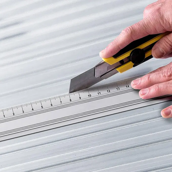 Floordirekt Doppelstegplatte Polycarbonat 60,5 x 121 cm 16 mm Transparent -  - Eine Hand schneidet mit einem gelben Cuttermesser entlang eines silbernen Lineals auf einer geriffelten Oberfläche.