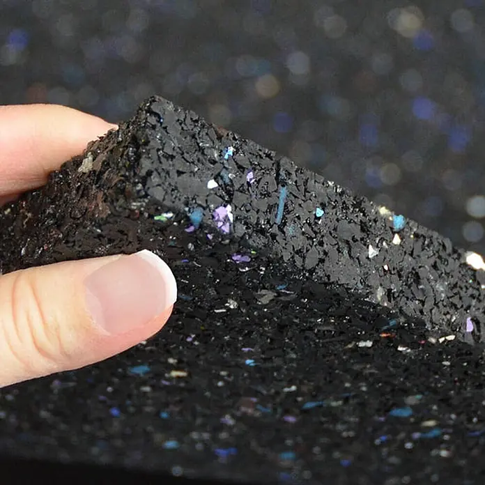 Floordirekt Antivibrationsmatte 60 x 60 x 1,97 cm -  - Hand hält eine dicke, flexible schwarze Matte mit bunten Sprenkeln, die ihre recycelte Gummigranulat-Textur und Dicke zeigt.