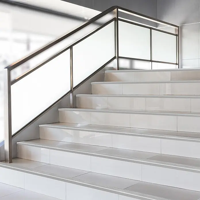 Floordirekt Stufenkantenprofil Safety F-Form Silber 90x5,3 cm Vorgebohrt -  - Modernes Treppenhaus mit weißen Fliesenstufen, grauen Antirutschstreifen und Metallgeländer mit Milchglasscheiben.