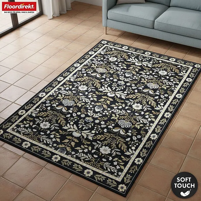 Floordirekt Kurzflorteppich Aristo 160 x 230 cm - Schwarz - Dunkler Teppich mit weißem und goldenem Blumenmuster, auf Fliesenboden vor einem hellblauen Sofa im Wohnzimmer.