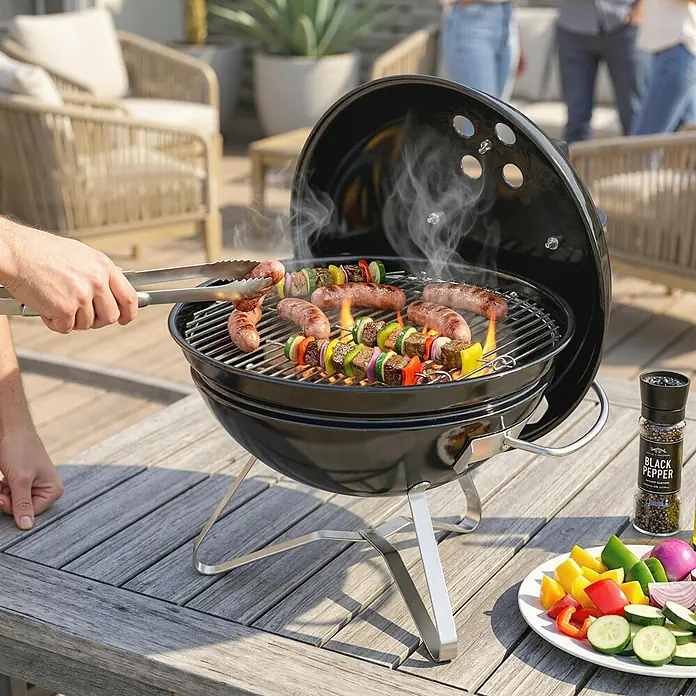 Outsunny-Kugelgrill-Ø41 x 44H cm -  - Schwarzer Holzkohlegrill mit Metallbeinen, der Würstchen und Gemüsespieße auf einem Holztisch im Freien brät.