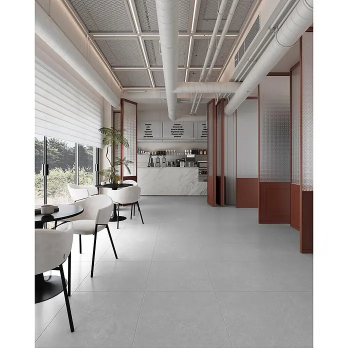 Feinsteinzeugfliese SOFT LIGHT GREY I.60 X – 60 x 60 cm, Steinoptik, Mat -  - Geräumiges, modernes Café-Interieur mit hellgrauen Fliesen, weißen Stühlen, schwarzen Tischen, Marmor-Theke, sichtbaren weißen Rohren und großen Fenstern.