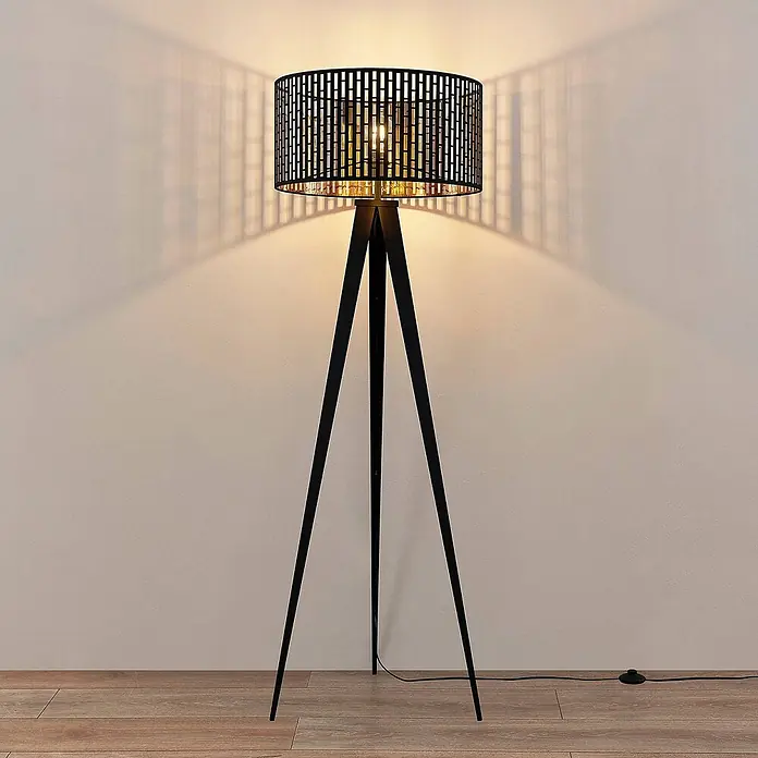 Lindby Stehlampe Dreibein schwarz gold mit Fußschalter, ohne Leuchtmittel -  - Schwarze Metall-Tripod-Stehleuchte mit gemustertem Zylinderschirm, die dekorative Lichtmuster an eine Wand wirft.