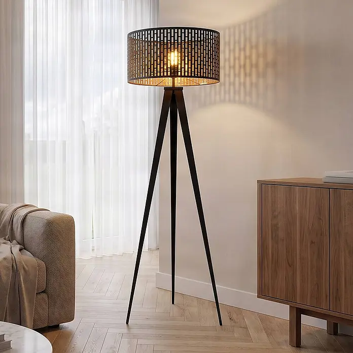 Lindby Stehlampe Dreibein schwarz gold mit Fußschalter, ohne Leuchtmittel -  - Schwarze Dreifuß-Stehlampe mit Metallgeflecht-Schirm, spendet Licht in einem Wohnzimmer.