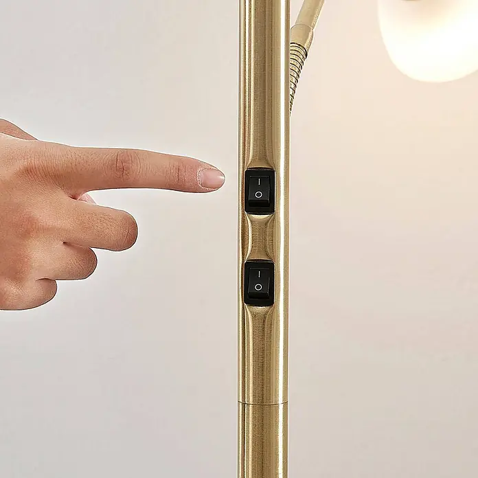 Lindby Stehlampe, Deckenfluter verstellbarer Lesearm, Standleuchte -  - Eine Hand bedient einen schwarzen Wippschalter an einer goldenen Messinglampe. Die Lampe hat zwei Schalter und einen flexiblen Arm.