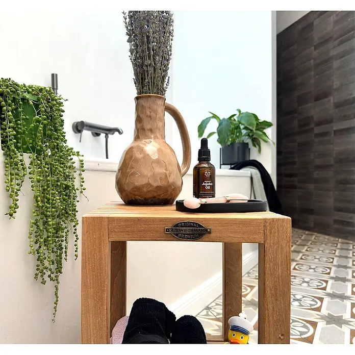 Badtisch Holz Leandro Teak massiv mit Gummifüßen -  - Hölzerner Hocker im Badezimmer mit Bio-Jojobaölflasche, pinkem Rosenquarz-Gesichtsroller und Lavendel in einer Vase.
