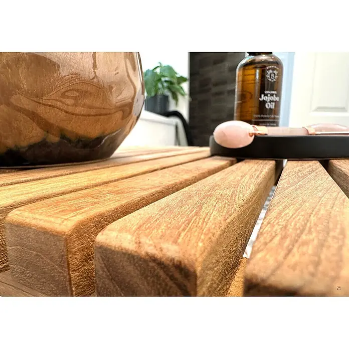 Badtisch Holz Leandro Teak massiv mit Gummifüßen -  - Eine bernsteinfarbene Jojobaölflasche und ein rosafarbener Gesichtsroller liegen auf einer Holzlamellenfläche.