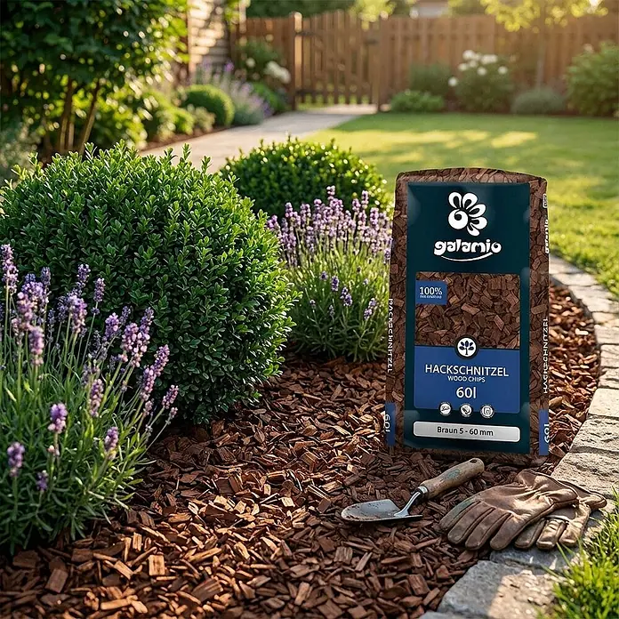 Hackschnitzel Holzdekor Braun 5-60 mm 60l -  - Tüte mit braunen Holzchips in einem Garten, als Mulch um Pflanzen verteilt, mit Gartengeräten daneben.