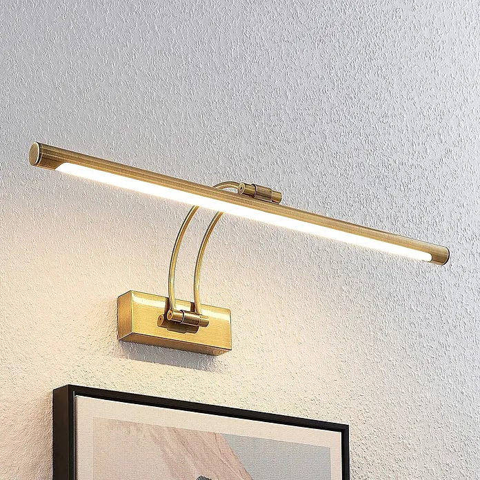 Lucande LED Wandlampe Bilderleuchte altmessing, warmweiß 3.000K -  - Messingfarbene Bilderleuchte an einer weißen Strukturwand montiert, die ein gerahmtes Kunstwerk beleuchtet.