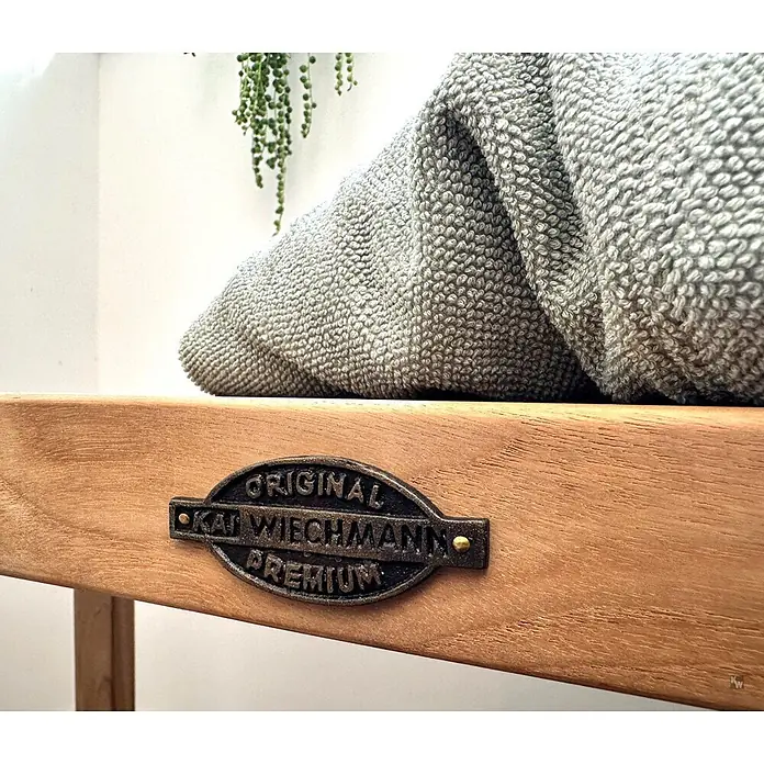 Bad Sitzbank Holz Belina als edle Teak Badezimmerbank massiv 75 x 31 cm -  - Holzbank mit dunkler Metallplakette 'ORIGINAL KAI WIECHMANN PREMIUM'. Eine gefaltete graue, strukturierte Decke liegt darauf.