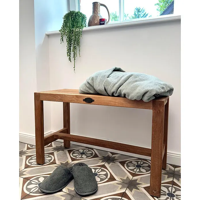 Bad Sitzbank Holz Belina als edle Teak Badezimmerbank massiv 75 x 31 cm -  - Eine Holzbank mit Lattenrost, darauf ein gefalteter hellgrüner Bademantel und ein Paar dunkelgrüne Hausschuhe auf gemustertem Boden.