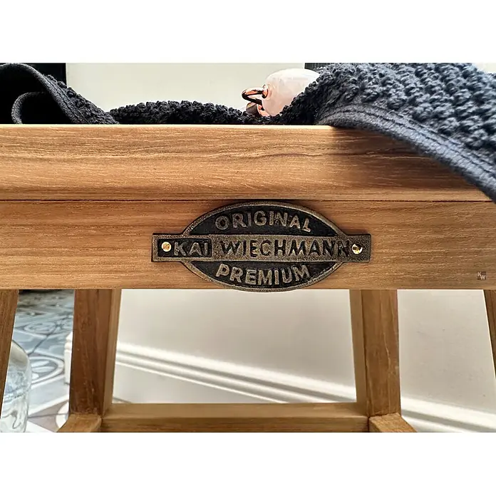 Bad Beistelltisch Holz Loretta Massivholz Hocker ohne sichtbare Schrauben -  - Holzmöbelstück mit dunklem Metallschild 'Kai Wiechmann Premium', das seine natürliche Holzmaserung in einem Wohnambiente zeigt.
