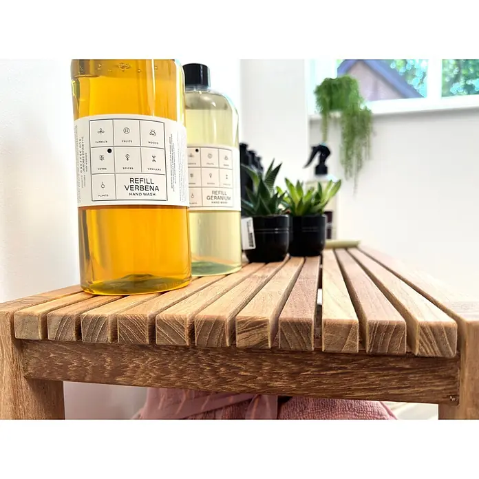 Badregal Holz Tito auch als Bad Sitzbank dank Massivholz-Konstruktion -  - Zwei bernsteinfarbene Nachfüllflaschen Handseife (Verbena, Geranium) mit weißen Etiketten auf einer hellbraunen Holzbank.