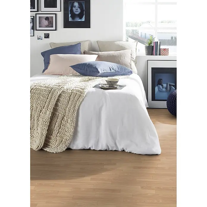 PVC-Boden Meterware Vinylboden 4m Schiffsboden Buche gedämpft -  - Bett mit weißer Bettdecke, verschiedenen Kissen (hellblau, rosa, beige) und einer cremefarbenen Grobstrickdecke, in hellem Schlafzimmer.