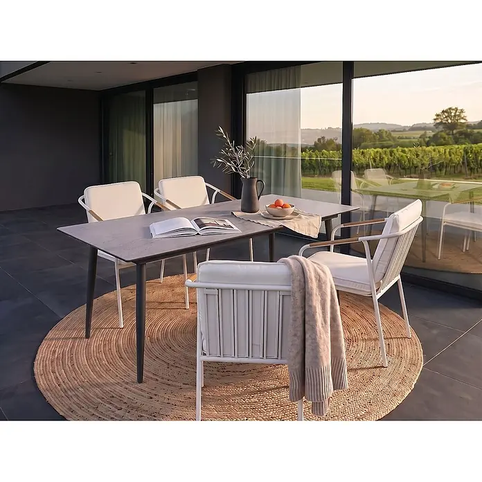 Esstisch TOLENTINO in Grau/Beige - (W) 90 x (H) 75 x - L - Outdoor-Essgruppe mit grauem Tisch, weißen Stühlen mit cremefarbenen Kissen und einem runden Jute-Teppich auf einer Terrasse.