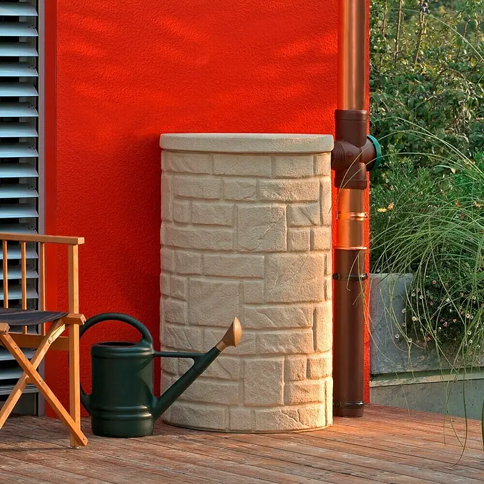Arves Regenspeicher Arcado - 230 l, Sandsteinfarben, 1 Stk. - Eine Regenwassertonne in Steinoptik steht auf einer Holzterrasse, neben einer grünen Gießkanne und einem Stuhl, an einer roten Wand.