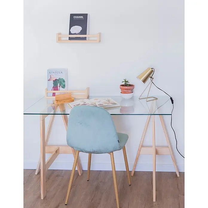 Astigarraga Encimera de cristal con caballetes - Vidrio templado, Pino, 120 x 60 cm - Un escritorio minimalista de cristal con patas de caballete de madera y una silla de terciopelo verde claro. Sobre la mesa hay una lámpara dorada, una planta y libros.