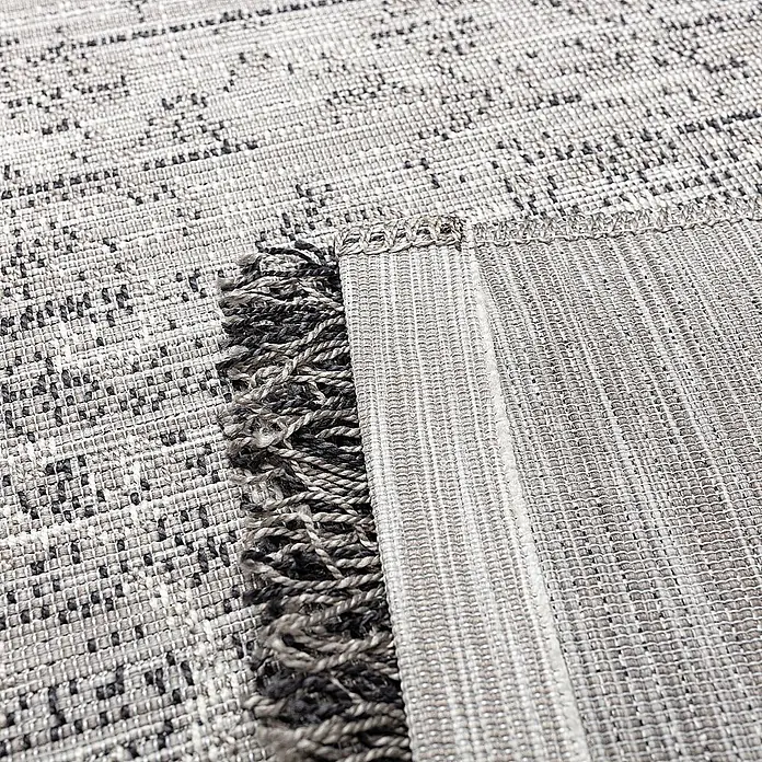 Dekoria Teppich Twed grey 120x170cm -  - Nahaufnahme eines gewebten grau-schwarz gemusterten Teppichs, gefaltet, um die strukturierte Unterseite und den Fransenrand zu zeigen.