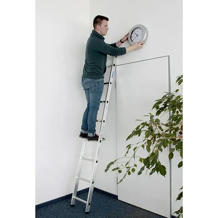 Krause Corda Escalera de apoyo - Número de peldaños: 8 ud., Altura de trabajo: 3,2 m, Carga soportada: 150 kg, Aluminio - Un hombre está subido a una escalera Krause y cuelga un reloj de pared redondo. Al fondo se ven una puerta y una planta de interior.