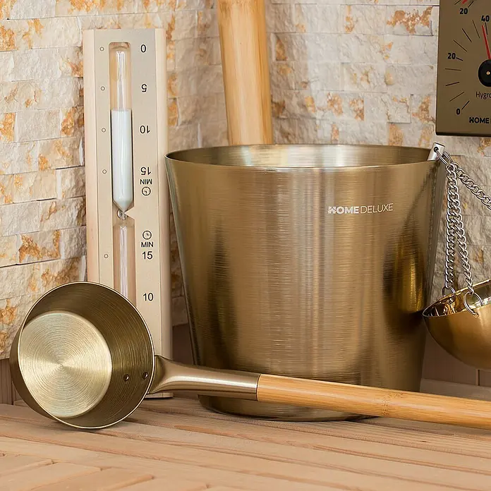 Home Deluxe Sauna-Zubehör THERA Gold -  - Goldener Saunaeimer, Schöpfkelle mit Bambusgriff und Sanduhr auf Holzsaunabänken vor einer Steinwand.