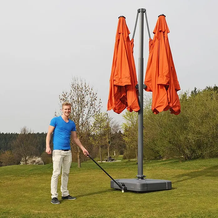 Mendler Gastronomie-Alu-Luxus-Doppel-Ampelschirm T44 - terracotta + Ständer -  - Großer orangefarbener Doppel-Ampelschirm, zusammengeklappt auf Rollfuß, von einem Mann bewegt.