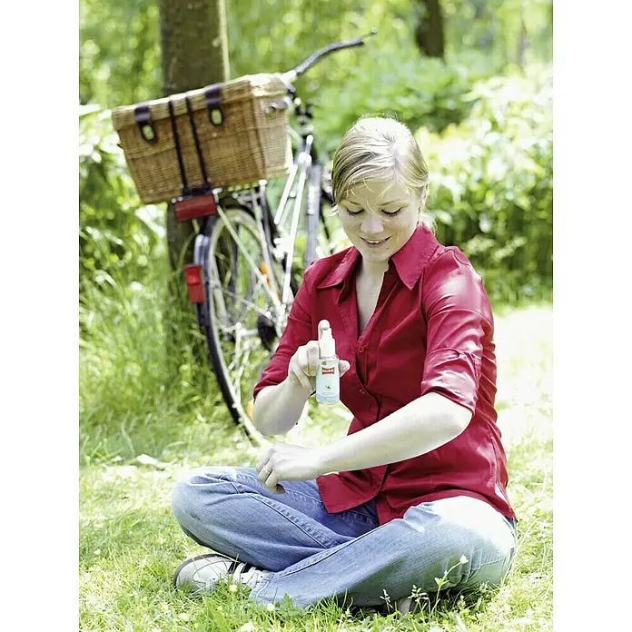 Ballistol Insektenschutzmittel Stichfrei - 100 ml - Frau trägt Insektenspray von BILLERBECK auf, sitzt auf einer Wiese mit Fahrrad im Hintergrund, idealer Mückenschutz für den Sommer.