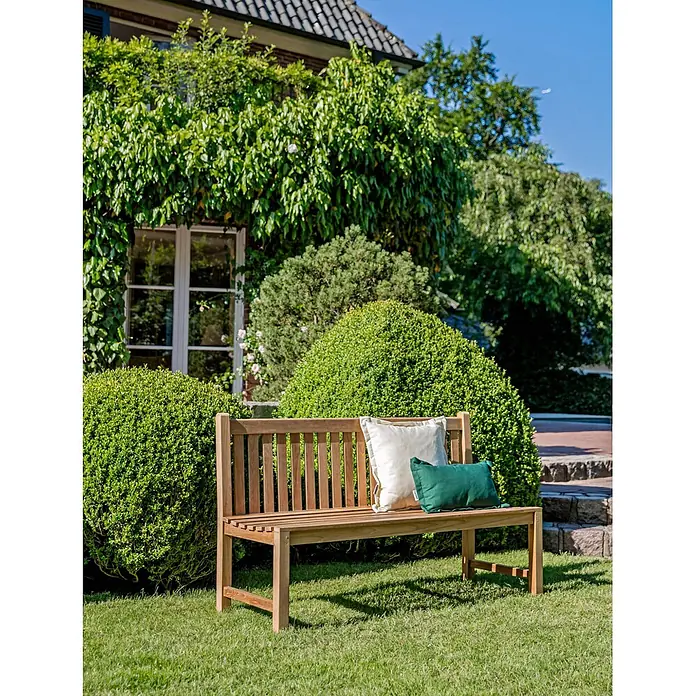 Teak Gartenbank ohne Armlehnen 110 cm wetterfest unbehandelt -  - Eine hellbraune Holzgartenbank mit einem weißen und einem grünen Kissen steht auf einem Rasen in einer üppigen Gartenumgebung.