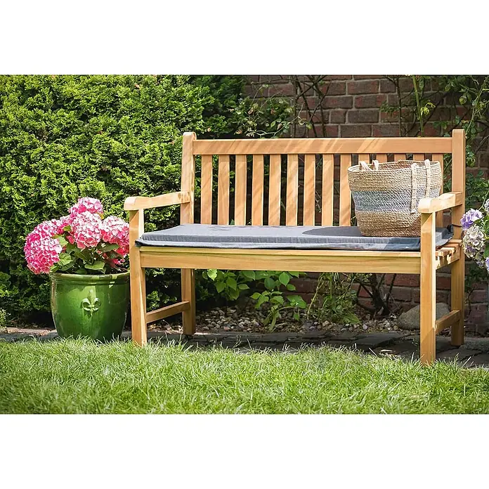 Teak Gartenbank 150 cm Holzbank massiv 3-Sitzer -  - Eine helle Holzgartenbank mit grauem Kissen steht in einem üppigen Garten neben rosa Hortensien, mit einem geflochtenen Korb darauf.
