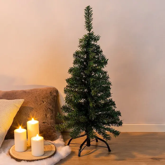 Künstlicher Weihnachtsbaum - Höhe: 90 cm, Grün - Ein grüner künstlicher Weihnachtsbaum steht in einem gemütlichen Raum mit angezündeten Kerzen auf einer Holzscheibe und weichen Kissen.