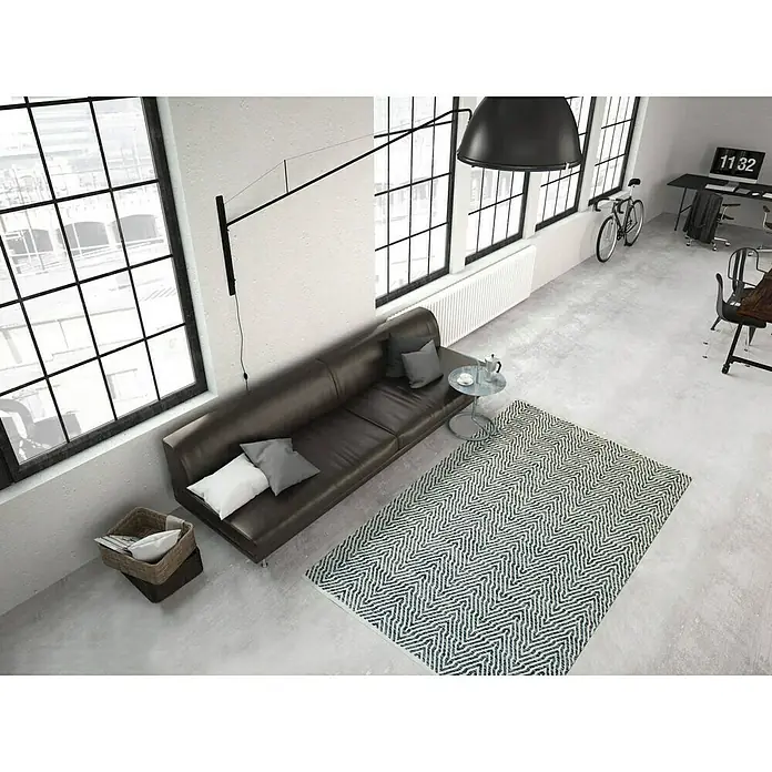 Kayoom Flachgewebeteppich Cocktail 400 - Grau, 170 x 120 cm, 100% Baumwolle - Eine stilvolle Wohnzimmerszene mit einem braunen Ledersofa, einem gemusterten Teppich und großen Fenstern, die viel natürliches Licht hereinlassen.