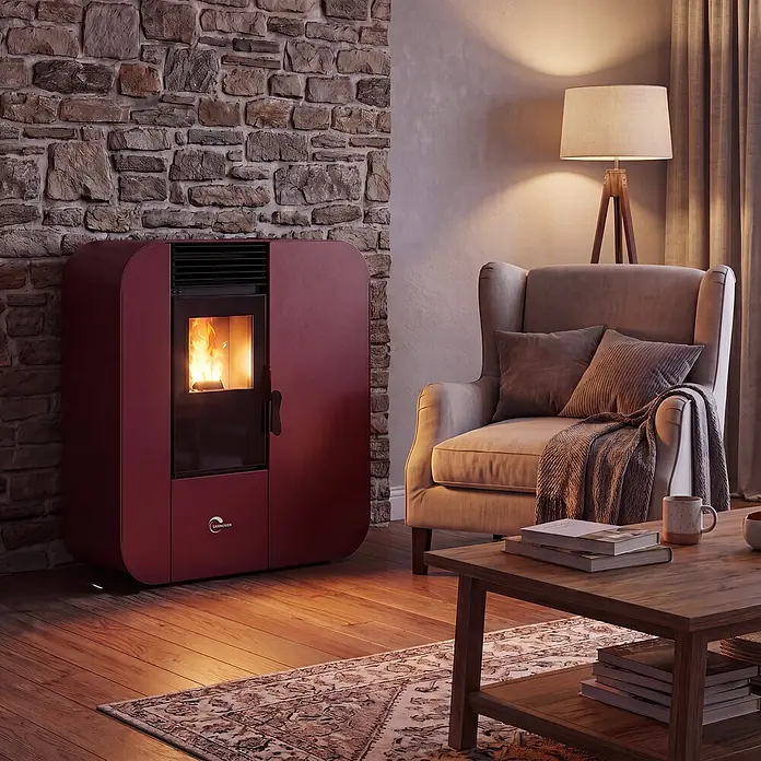 Pelletofen Slim Astral 13,8 kW Bordeaux Sannover -  - Roter Pelletofen mit sichtbarer Flamme, der ein gemütliches Wohnzimmer neben einer Steinwand und einem Sessel wärmt.