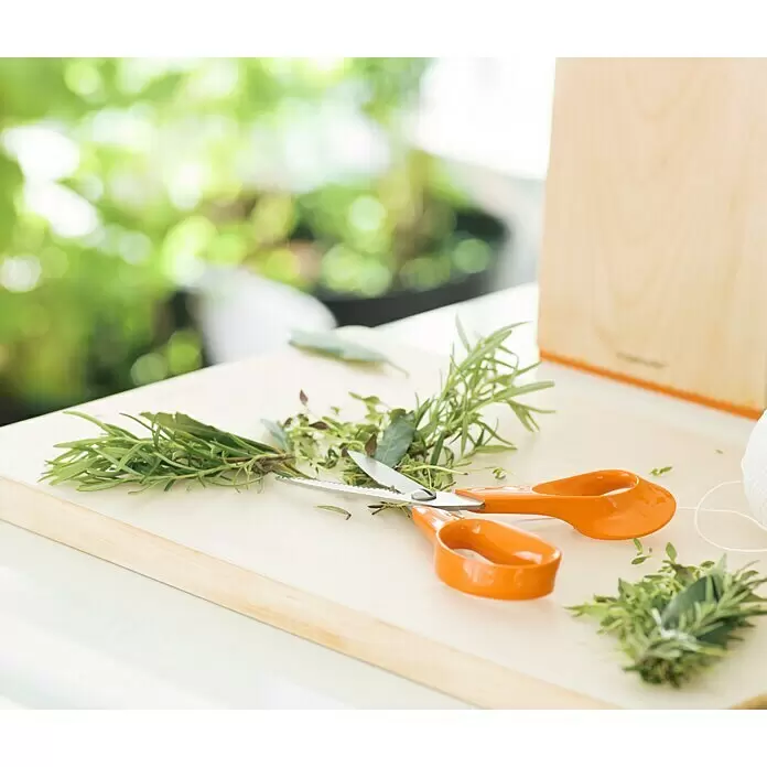 Fiskars Tijera universal - Largo: 18 cm, Acero - Tijeras de cocina naranjas, hierbas frescas y cordel blanco sobre una tabla de madera clara.