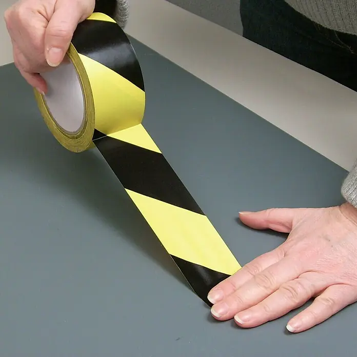 Cinta de señalización adhesiva - Amarillo/Negro - Una persona aplica un rollo de cinta de peligro a rayas amarillas y negras sobre una superficie gris. La cinta de peligro se utiliza para marcar zonas peligrosas.