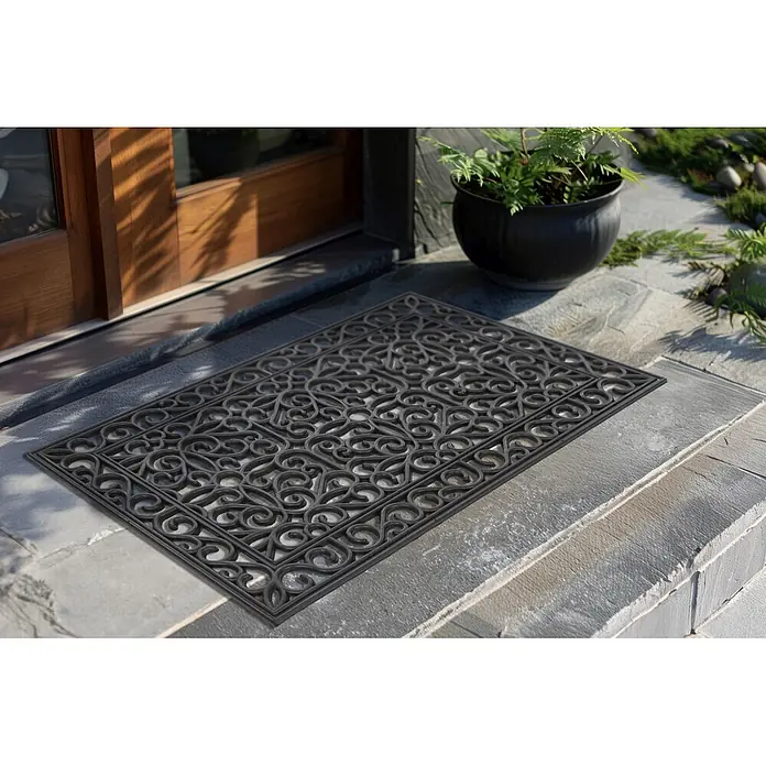 Fußmatte Everyday 125 - 75 x 45 cm, Schwarz, Material Nutzschicht: 100 % Gummi - Dunkle, verzierte Gummifußmatte mit kunstvollem Schnörkelmuster, auf einer Steinstufe vor einer Holztür platziert.